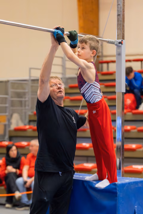 An older coach steadies a focused young boy gripping the high bar during a gymnastics competition in a red-seated arena.