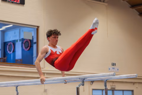 Young male gymnast holds a straddle L-sit on parallel bars, legs raised high, expression intense and focused.
