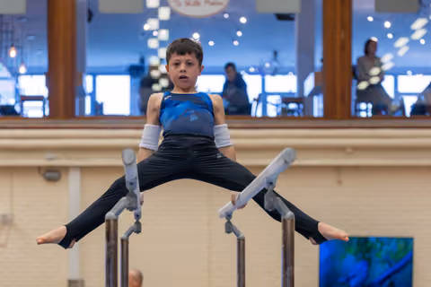 Young boy performs a straddle hold on parallel bars, legs spread wide, expression serious and concentrated in a gymnasium.