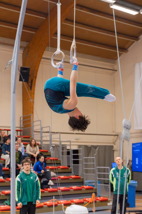 Young male gymnast hangs inverted on still rings, body arched, coaches watching below in indoor sports hall.