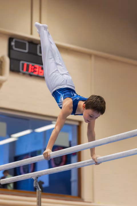 Young boy performs a handstand on parallel bars, legs split wide, face showing intense concentration in an indoor gym.