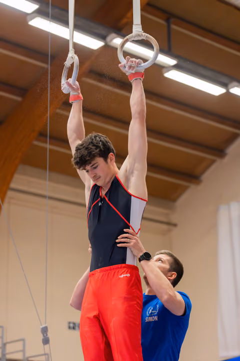 Young male gymnast grips still rings with intense focus while coach Simon supports his waist in an indoor gym.