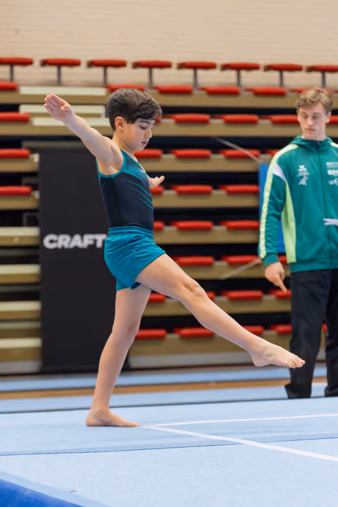 Young gymnast performs a focused floor routine, leg extended mid-step, while a coach in green watches attentively behind.