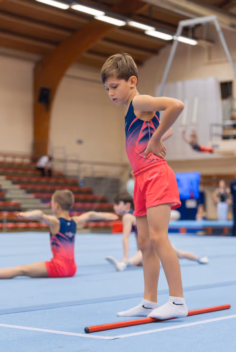 Young boy in red and navy leotard stands focused on a floor rod, hands on hips, in a gymnastics hall.