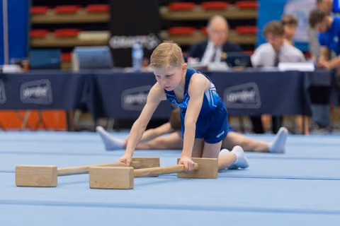 Focused young boy in blue leotard performs floor exercise with wooden apparatus blocks at a gymnastics meet.