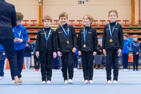 Four young boys in black tracksuits proudly display gold medals at a gymnastics competition, smiling on the competition floor.