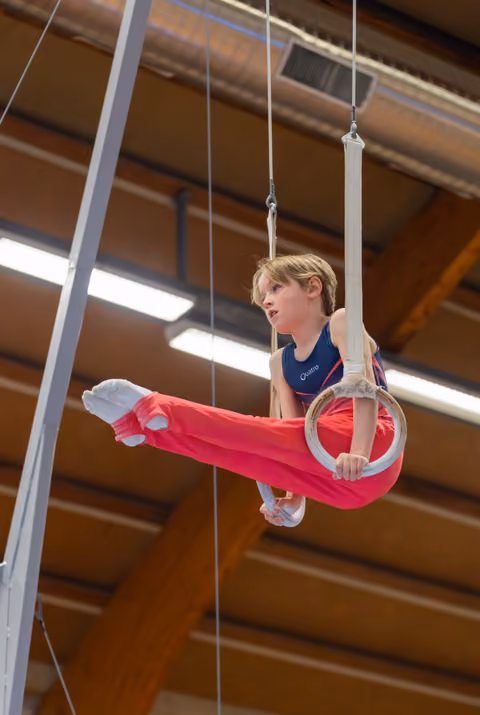 Focused young boy performs a straddle hold on gymnastics rings in an indoor sports hall, legs extended with intense concentration.