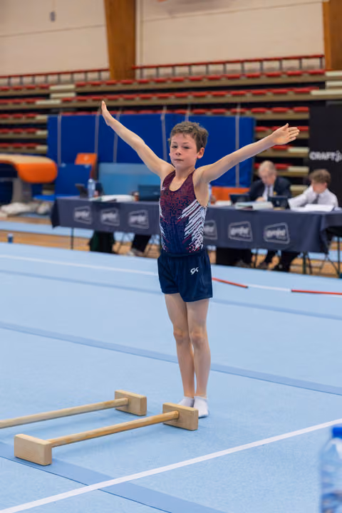 Young boy gymnast stands proudly with arms outstretched on blue floor mat, judges panel visible behind him.