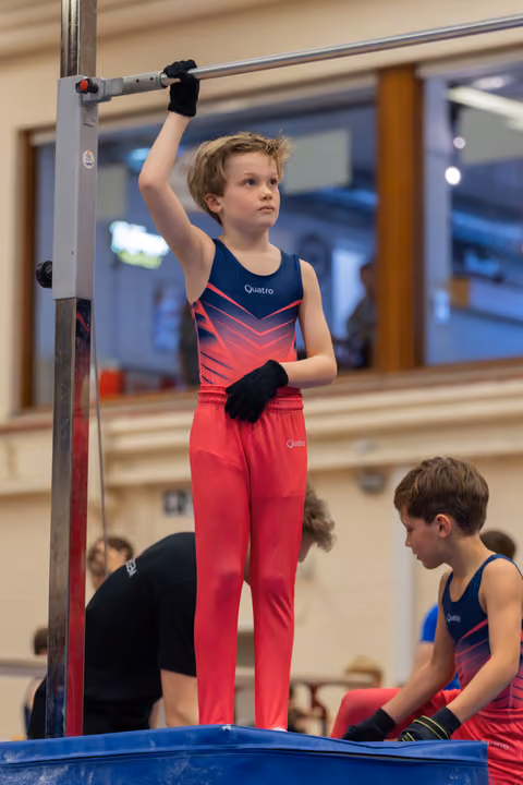 Focused boy in red Quatro leotard grips the high bar, standing tall, while a teammate waits nearby in a gym.