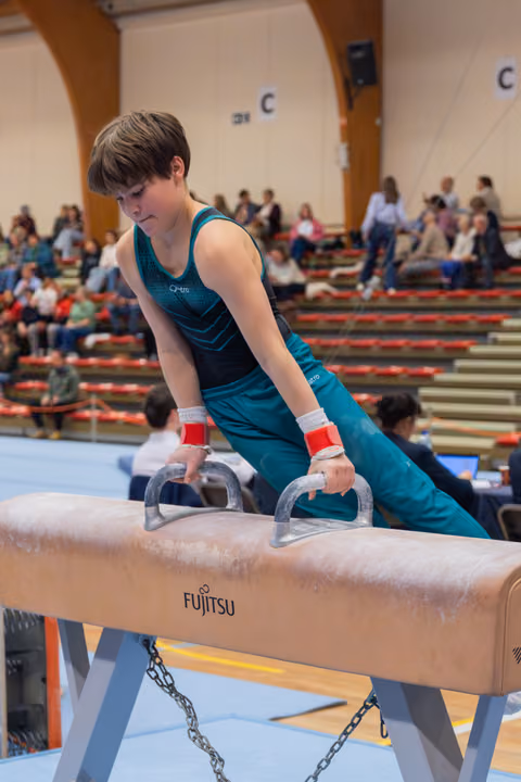 Focused young male gymnast grips pommel horse handles, leaning forward with intense concentration during a gymnastics competition.
