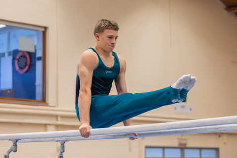 Young male gymnast performs a straddle hold on parallel bars, focused expression, teal leotard, indoor gym.