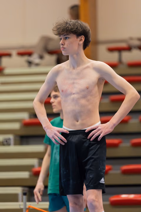 Shirtless teen gymnast stands with hands on hips, focused expression, in a gymnasium with red bleachers behind.