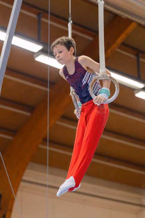 Focused young boy grips gymnastic rings mid-routine, body angled with concentration in a wooden-beamed sports hall.