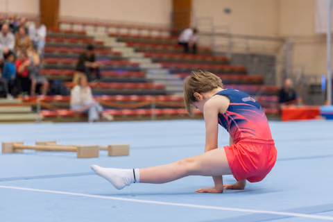 Young gymnast in red and navy leotard performs a split stretch on the blue floor mat, head bowed in focus.