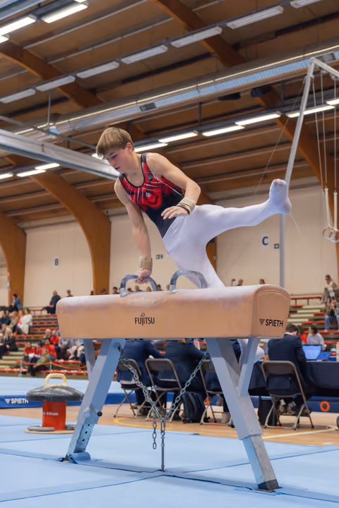 Young male gymnast performs on pommel horse, leaning forward with intense concentration, legs split mid-routine in indoor arena.
