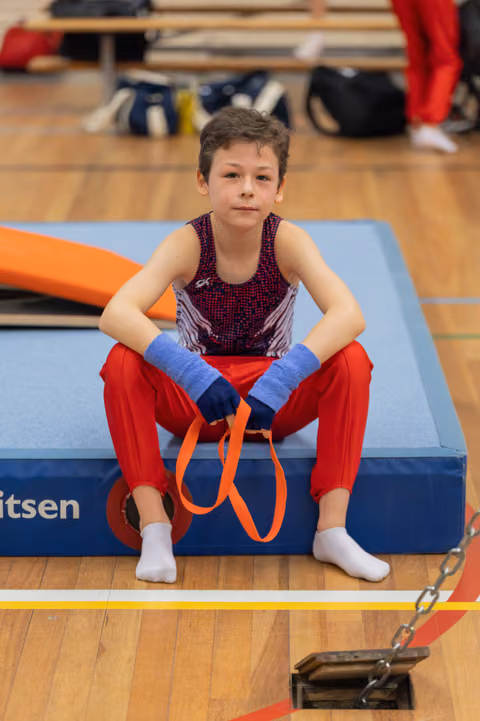 Young male gymnast sits on a blue mat holding orange straps, looking calm and focused between routines in a gym.