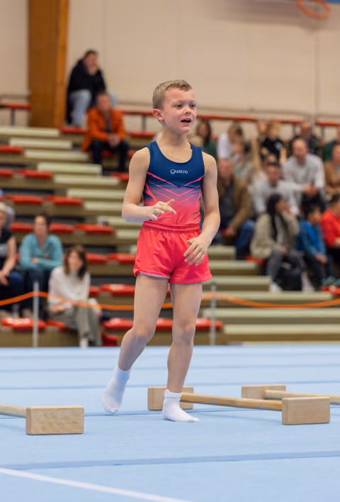 Young boy in blue and pink Quatro leotard performs floor routine, expression intense, audience watching in bleachers behind.