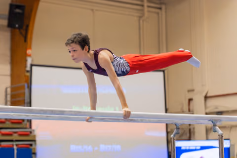 Young male gymnast holds a planche on parallel bars, body horizontal, expression focused and determined during a gymnastics meet.