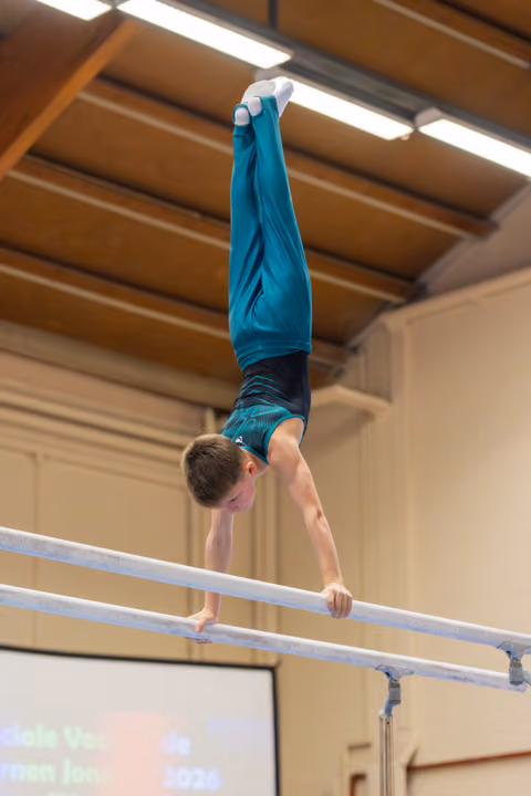 Young male gymnast performs a perfect handstand on parallel bars, legs extended upward, in an indoor gymnastics hall.