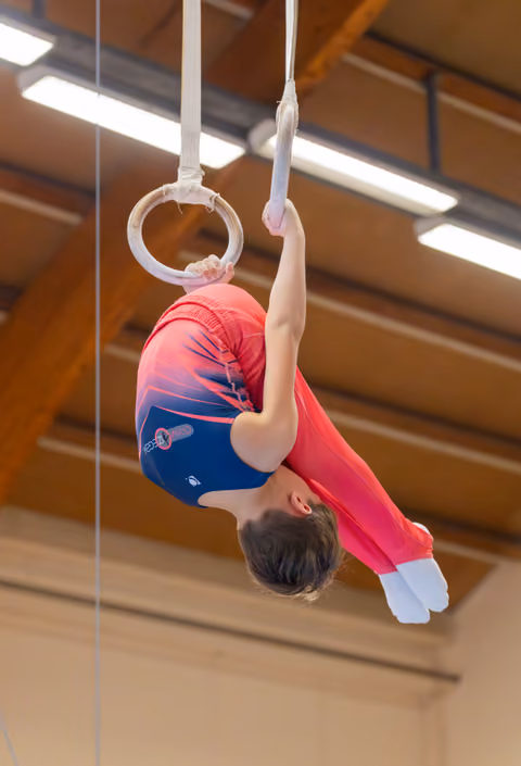 Young male gymnast hangs inverted on still rings, body arched, wearing red and blue leotard in a wooden-beamed gymnasium.