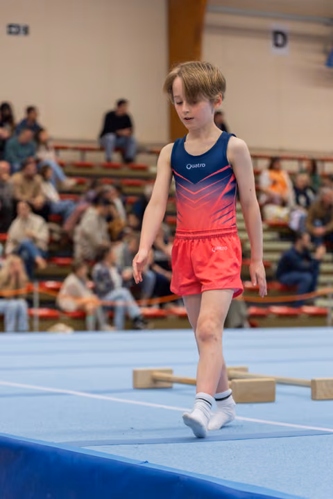 Focused young boy in red and navy Quatro leotard walks carefully across a blue gymnastics mat before a crowd.
