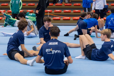 Young male gymnasts in navy GO4gym shirts sit on the floor mat laughing and chatting together between events.