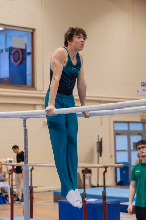 Focused young male gymnast balances on parallel bars, arms taut, looking ahead with concentration in an indoor gymnasium.