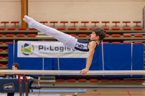 Young male gymnast holds a straddle position on parallel bars, body horizontal, focused expression in an indoor gymnasium.