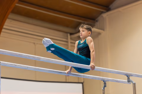 Focused young boy performing a leg raise on parallel bars, wearing a teal leotard, in an indoor gymnastics hall.