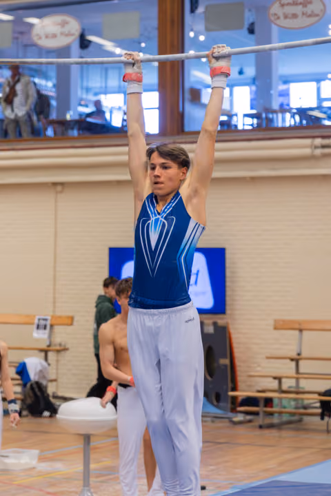 Young male gymnast hangs from the high bar, focused expression, wearing blue leotard and white pants in an indoor gym.