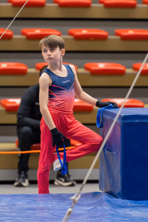 Young male gymnast in red and navy Quatro leotard rests a leg on a blue vault, looking focused and composed.