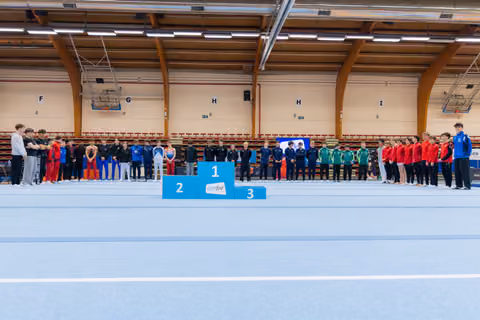 Teams line up around a 1-2-3 podium at an indoor gymnastics meet, awaiting a medal ceremony with calm anticipation.