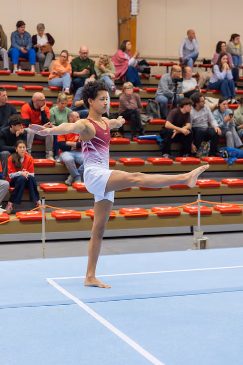 Young male gymnast holds a confident arabesque balance on the floor mat, arms extended, leg raised high.