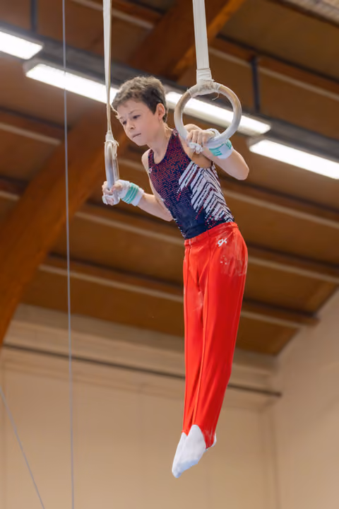 Young male gymnast hangs from still rings with intense focus, body angled, wearing red pants and sequined leotard indoors.