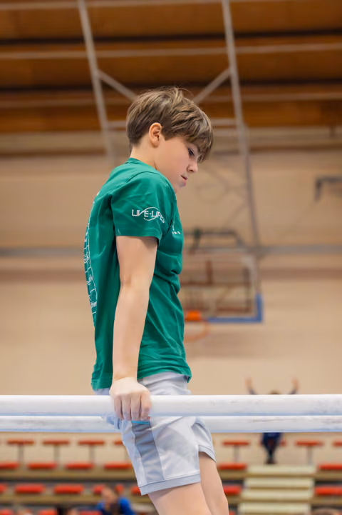 Young boy in green t-shirt leans on parallel bar, looking down with focused concentration in a gymnasium.