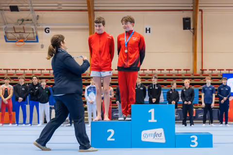 Woman pins a medal on a smiling boy in red on the 1st place podium, as his teammate stands on 2nd.