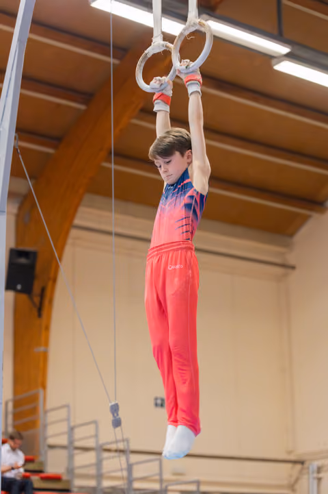 Focused young male gymnast hangs from still rings, looking down with concentration, in a wooden-beamed sports hall.