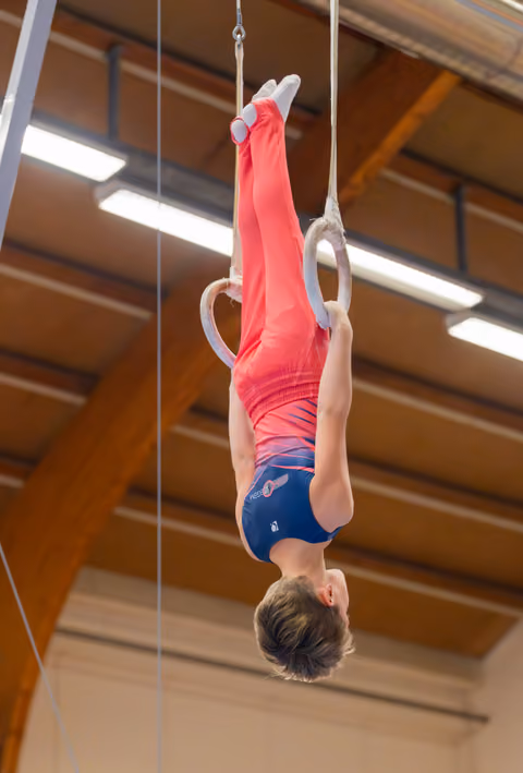 Young male gymnast performs an inverted hang on still rings, body fully extended, in an indoor gymnastics hall.