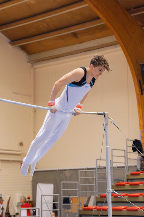 Focused teenage gymnast grips the high bar, body horizontal, displaying strength and concentration in an indoor gymnastics hall.