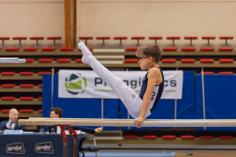 Young boy in glasses performs a precise leg raise on parallel bars, showing intense focus during a gymnastics meet.