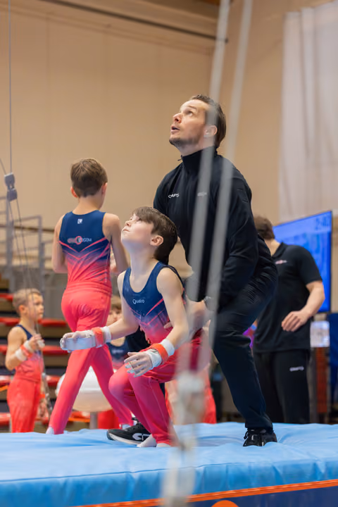 Coach spots young gymnast on mat, both gazing upward intently during a youth gymnastics meet.