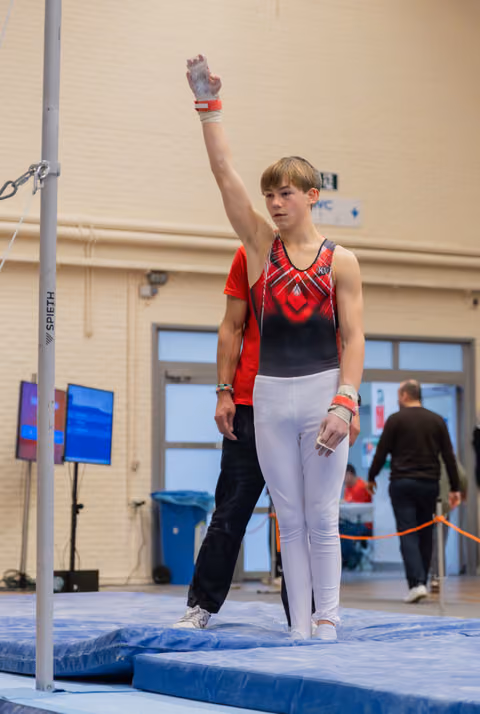 Young male gymnast raises one hand in salute before the high bar, composed and focused, coach standing behind him.