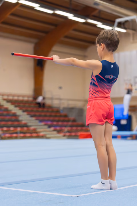 Young boy in red and navy leotard stands poised on blue mat, extending a red stick outward with focused posture.