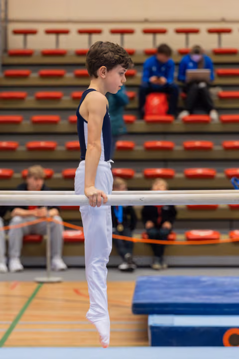 Young male gymnast grips parallel bars with focused concentration, wearing navy leotard and white trousers in a gymnasium.