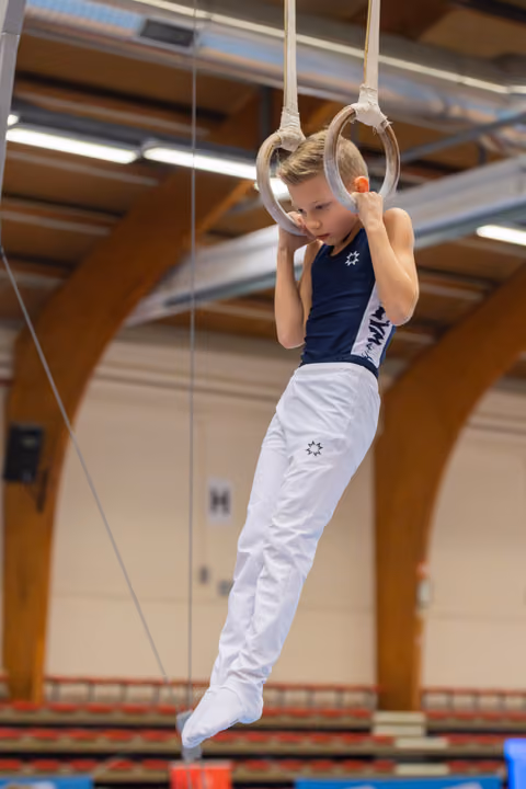Focused young male gymnast grips still rings mid-routine, legs tucked, intense concentration in a gymnasium.