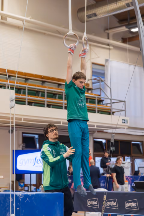 Young gymnast hangs from still rings while his coach watches attentively with hands clasped at a Gymfed competition.