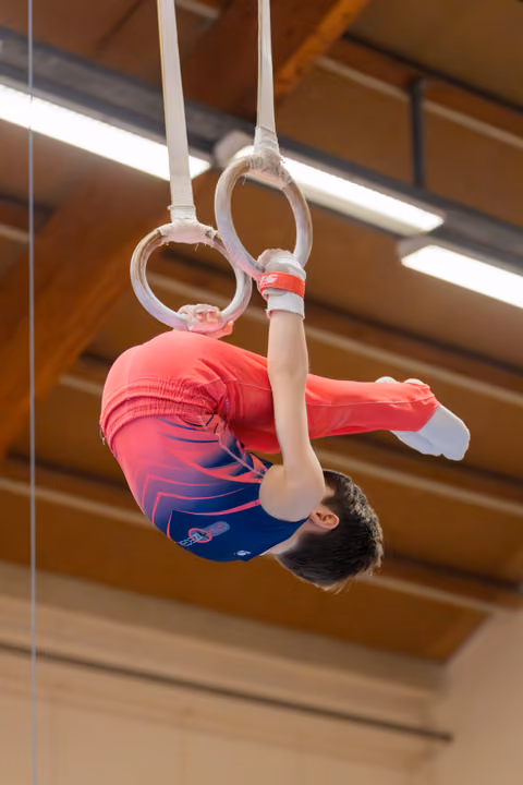 Young male gymnast holds an inverted position on still rings, face calm and focused, in a wood-panelled gymnasium.