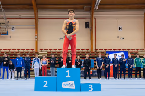 Young male gymnast stands proudly on the first-place podium at a gymfed indoor gymnastics championship, arms crossed.