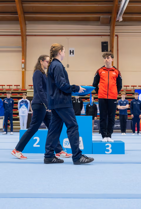 Two officials in navy tracksuits present a medal to a young gymnast in red standing on the third-place podium, expression serious.