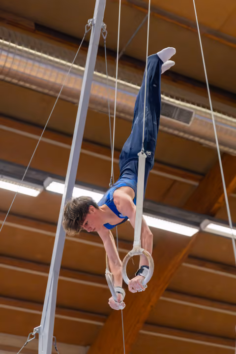 Young male gymnast performs an inverted pike hold on still rings, showing intense focus and strength in an indoor gymnasium.